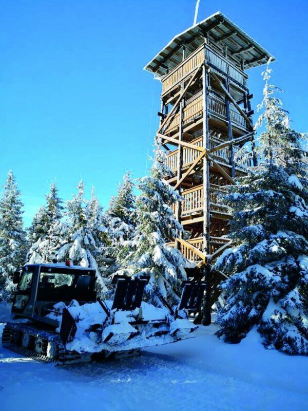 Widok na drewnianą wieżę widokową w otoczeniu ośnieżonych drzew z widocznym przejeżdżającym ratrakiem na pierwszym planie. Słoneczna pogoda.