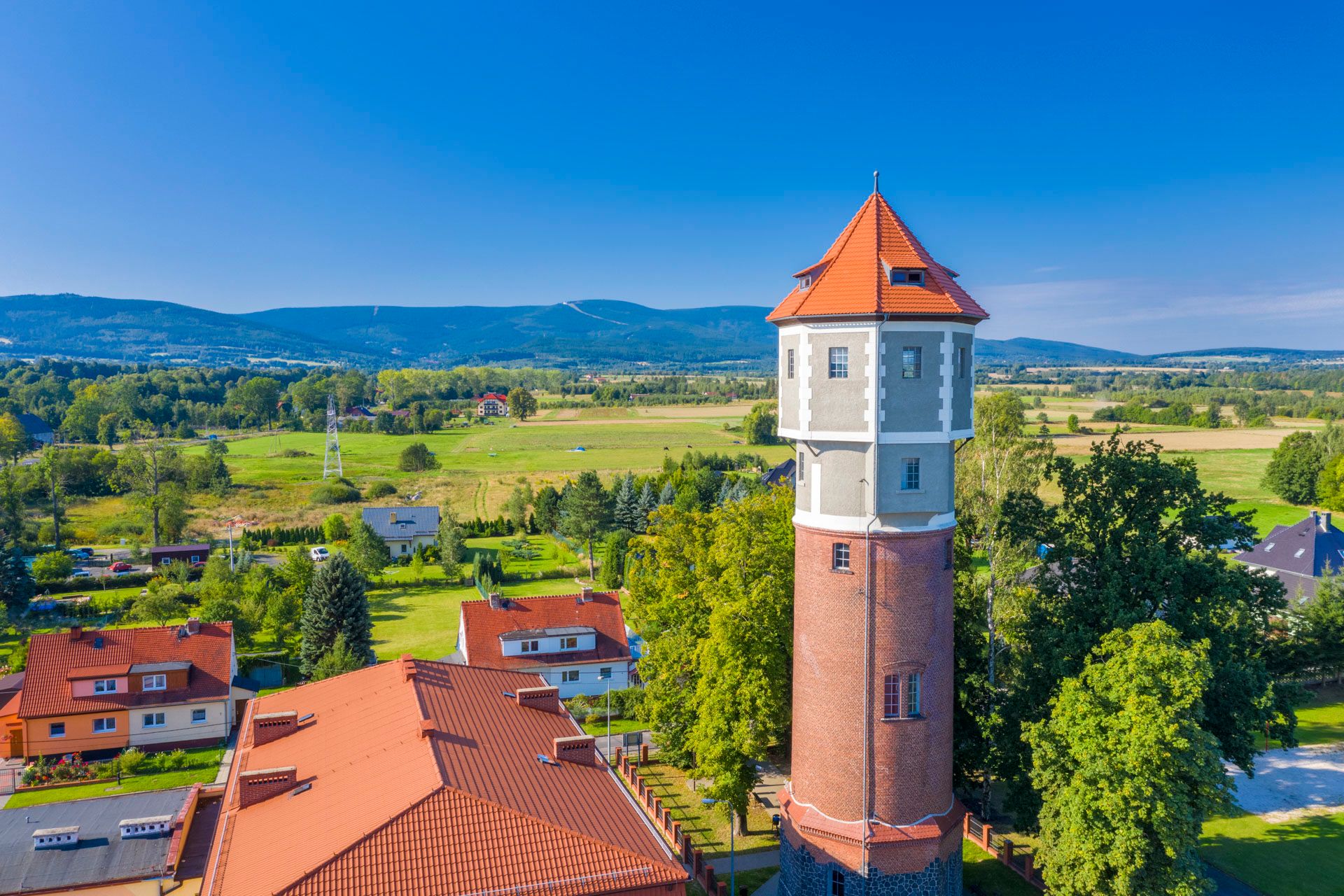 Zdjęcie z drona przedstawiające ceglaną wieżę ciśnień z odciętym szczytem w kolorze grafitowym na tle zieleni oraz błękitu nieba. 