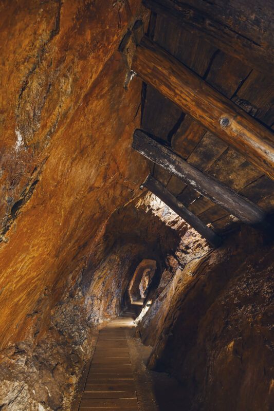 Lekko mroczny tunel wydrążony w skale. Na końcu korytarza lekkie światło, miejscami drewniane elementy oraz podłoga.