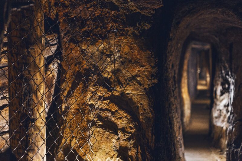 Lekko mroczny tunel wydrążony w skale. Na końcu korytarza lekkie światło, miejscami drewniane elementy i siatka.