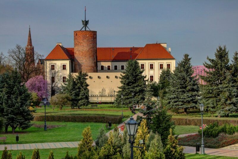 Widok na zabytkowy budynek muzeum archeologicznego z widoczną ceglaną wieżą oraz kwitnącym, zadbanym ogrodem z licznymi ścieżkami na pierwszym planie.
