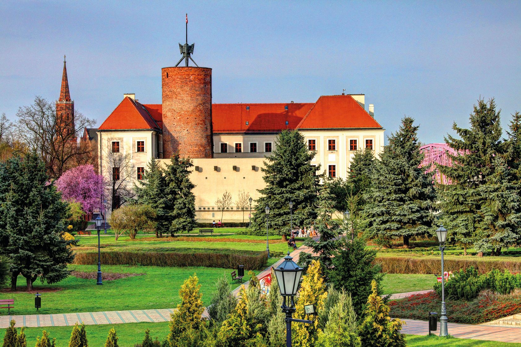 Widok na zabytkowy budynek muzeum archeologicznego z widoczną ceglaną wieżą oraz kwitnącym, zadbanym ogrodem z licznymi ścieżkami na pierwszym planie.