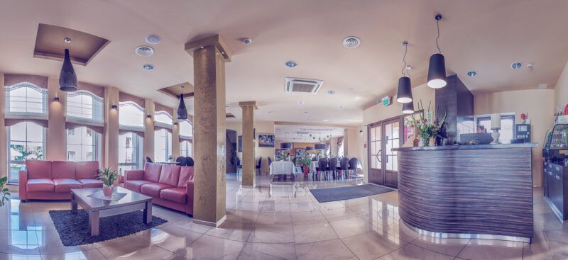 Recepcja hotelowa wraz z pomieszczeniem ogólnodostępnym. Na zdjęciu widać kanapy do siedzenia kolumny w pomieszczeniu oraz duże okna wpuszczające wiele światła do środka