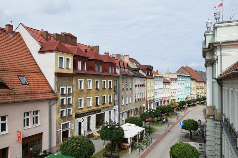 Dwa rzędy różnokolorowych kamienic. Na środku, pomiędzy nimi ogródki restauracyjne, elementy zieleni miejskiej.