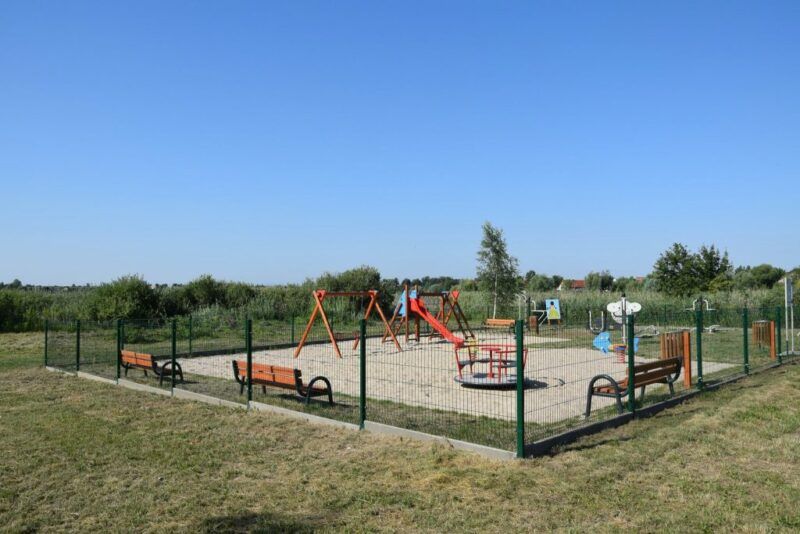 Plac zabaw dla dzieci z huśtawką, minikaruzelą oraz ławkami i ogrodzeniem.