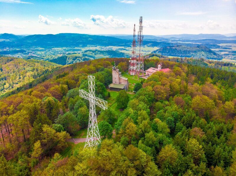 Metalowy krzyż oraz wieża radiowo-telewizyjna na szczycie góry, porośniętej zielonym lasem.