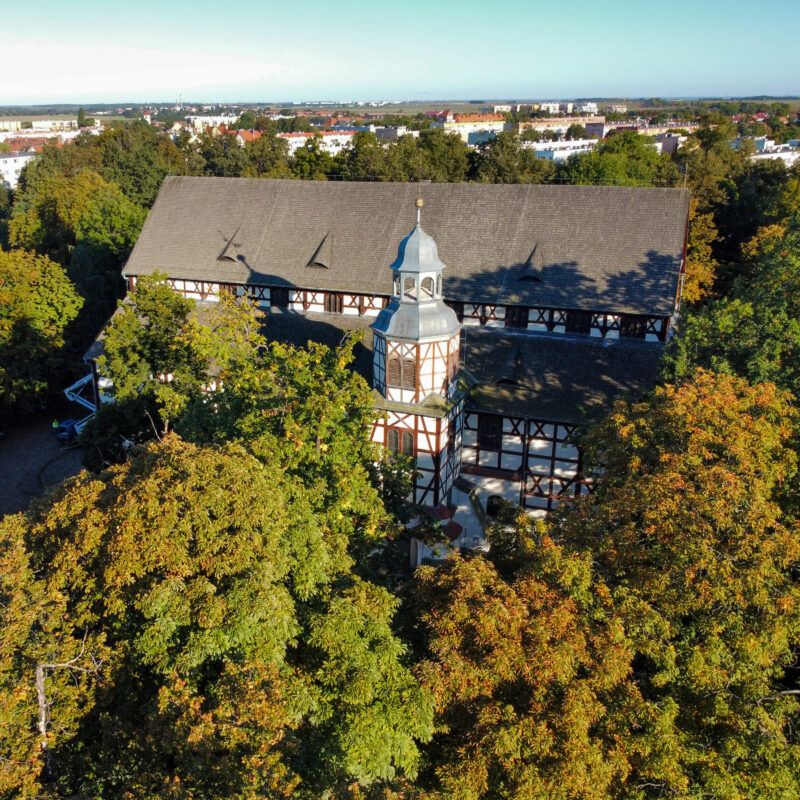 Zdjęcie z drona przedstawiające biało-brązową bryłę kościoła szachulcowego, wynurzającą się spośród drzew w parku, 