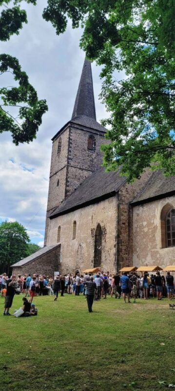 Romański kościół zbudowany z jasnego kamienia. Przed nim sporo ludzi biorących udział w festynie.