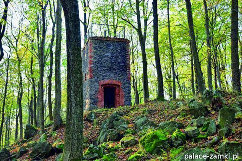 Widok na niską, kamienną wieżę widokową z ceglanymi narożnikami położoną na zboczu góry w otoczeniu drzew