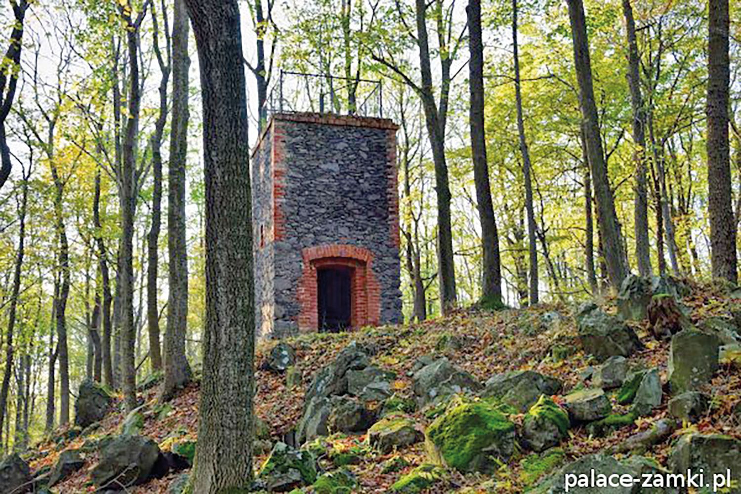 Widok na niską, kamienną wieżę widokową z ceglanymi narożnikami położoną na zboczu góry w otoczeniu drzew