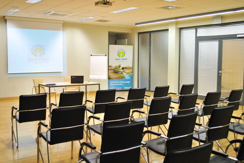 Sala konferencyjna z ustawionymi równo krzesłami, zwróconymi w stronę ekranu. Po prawej stronie został ulokowany stojak z reklamą obiektu Aqua-Zdrój.