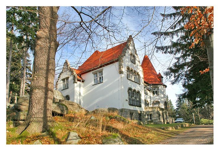 stara biała willa z czerwonym dachem w otoczeniu drzew nad którą rozpościera się jasne niebo w kolorze błękitu.