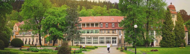 Zabytkowy, ogromny budynek uzdrowiska Jan Kazimierz w otoczeniu zieleni.