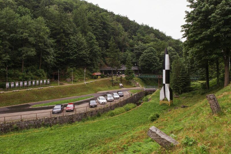 Widok na parking, na którym stoją zaparkowane samochody w otoczeniu zieleni i otulinie drzew. Na drugim planie widoczne ciemne budynki kompleksu.