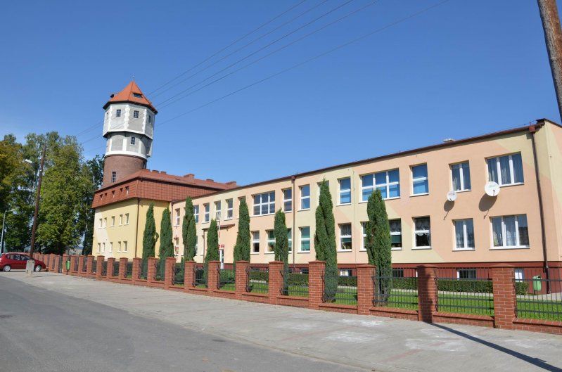 Ceglana wieża ciśnień z odciętym szczytem w kolorze grafitowym oraz sąsiadujący z nią podłużny budynek 