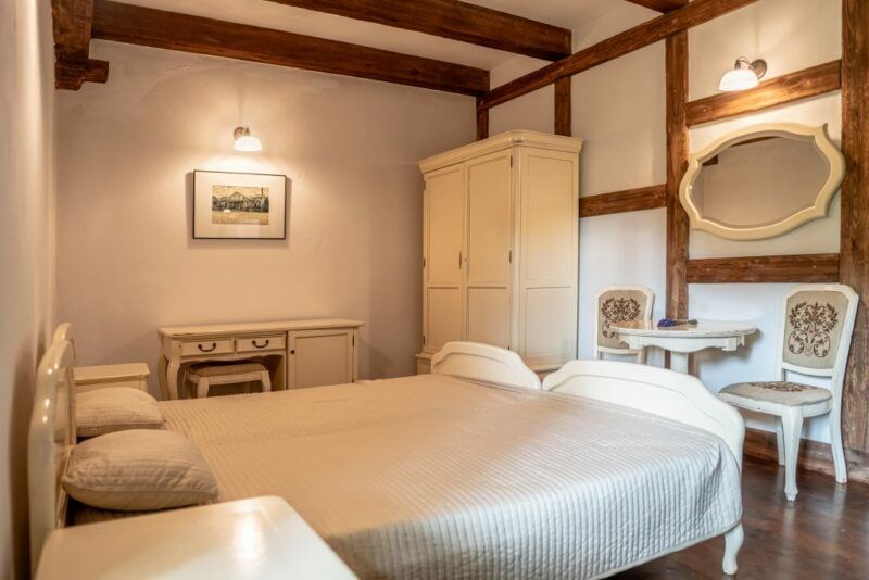 Pokój hotelowy w barwach białych, w którym jest lustro, łóżko, szafa stół, krzesła. W pokoju święcą się lampy naścienne w ciepłym odcieniu.