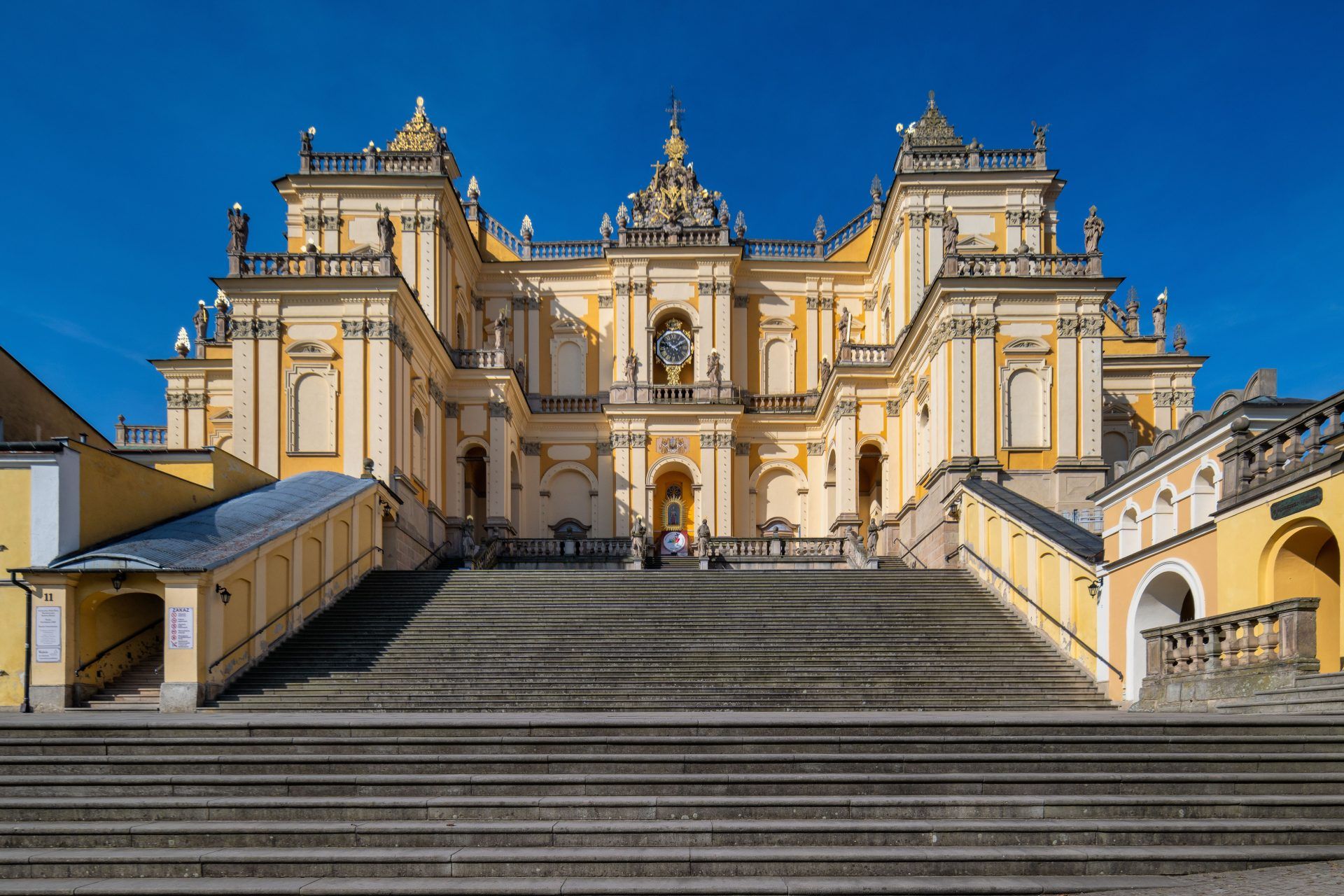 Na zdjęciu widać okazałe sanktuarium w Wambierzycach z monumentalnymi schodami prowadzącymi do barokowej fasady ozdobionej kolumnami i wieżami.