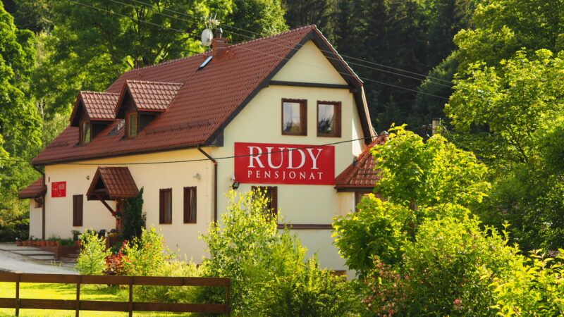 Trójkondygnacyjny budynek pensjonatu z szyldem z nazwą Rudy, skąpany w letnim słońcu na tle bujnej zieleni lasu.