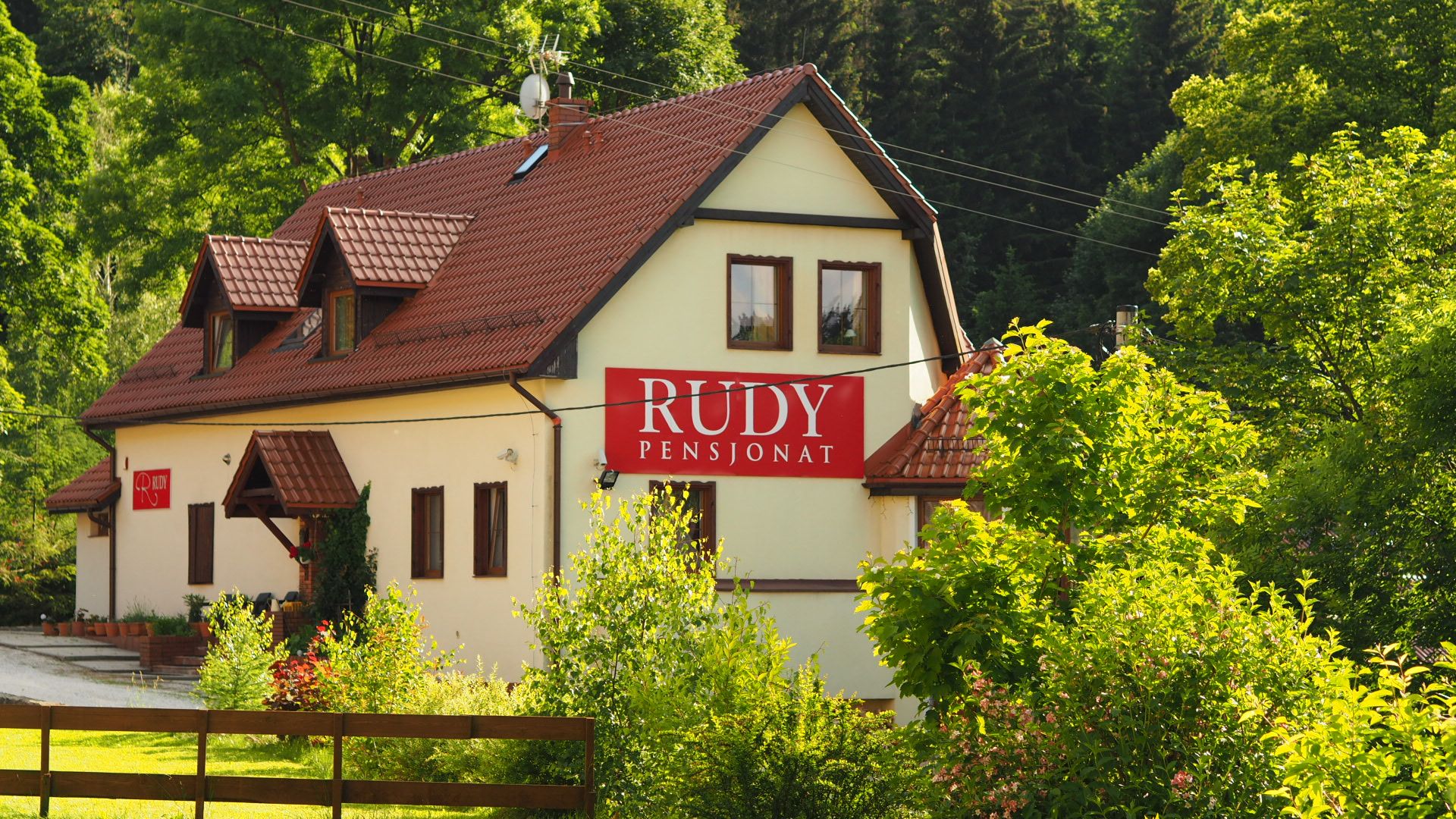 Trójkondygnacyjny budynek pensjonatu z szyldem z nazwą Rudy, skąpany w letnim słońcu na tle bujnej zieleni lasu.