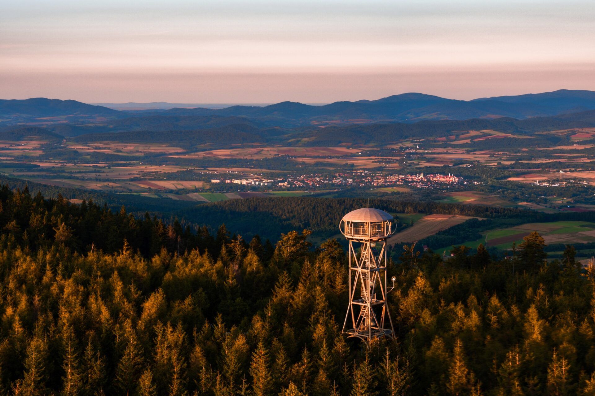 Widok z lotu ptaka na wieżę widokową, wzgórza, lasy oraz miasto w oddali.