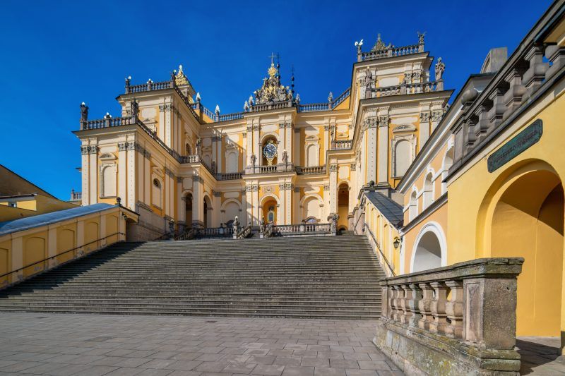 Na zdjęciu widać okazałe sanktuarium w Wambierzycach z monumentalnymi schodami prowadzącymi do barokowej fasady ozdobionej kolumnami i wieżami.