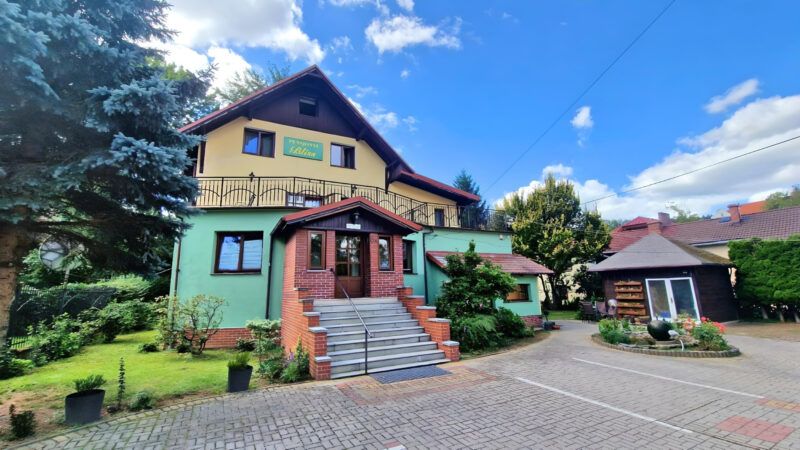Dwukondygnacyjny budynek z rozległym tarasem, ze spiczastym, stromym dachem i murowanym gankiem na wejściu.