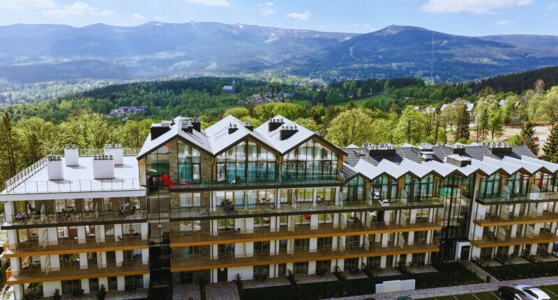 Widok na duży, wielopiętrowy budynek hotelowy na tle gór, w letnim słońcu. 