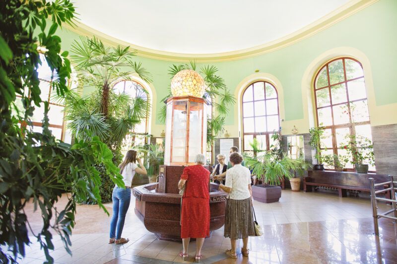 Wnętrze zabytkowego budynku, w którym znajduje się dostęp do uzdrowiskowej wody. w centralnej części zdjęcia stoją ludzie korzystający ze źródła w tle widać duże stare okna i ściany w kolorze pastelowo zielonym 