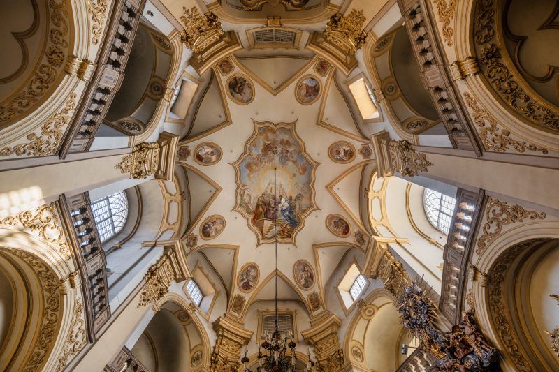 Na zdjęciu widać ozdobny, barokowy sufit sanktuarium w Wambierzycach, pokryty malowidłami i złoceniami w jasnych, ciepłych tonach.