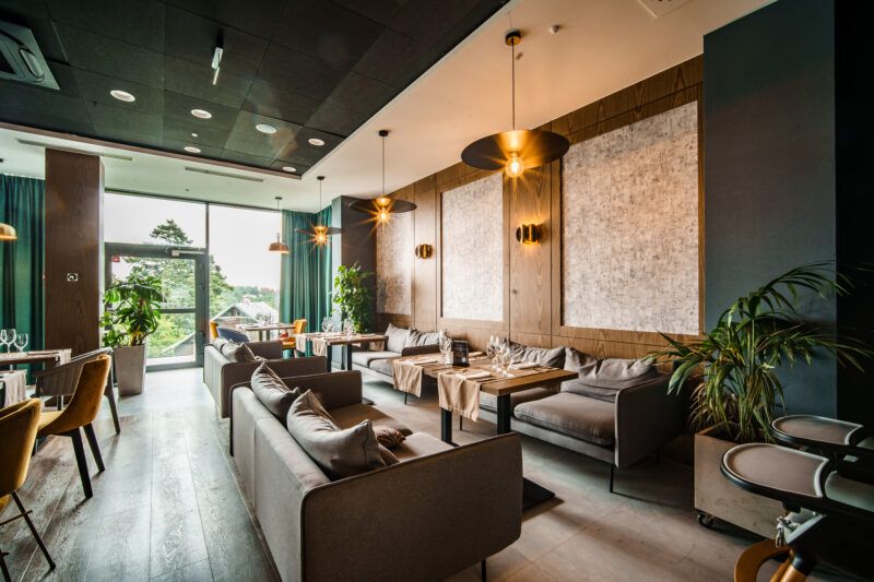 Restauracja hotelowa. Kilkanaście dużych kanap wypoczynkowych z drewnianymi stolikami. Duże przeszklone okno, na ścianach drewniane panele.