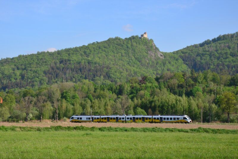 Widok na górę obrośniętą drzewami, pod górą przejeżdża pociąg Koleje Dolnośląskie.