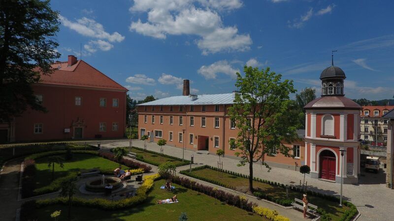 Widok na uzdrowisko, różowy budynek, przed nim kilka drzew, zadbany trawnik.