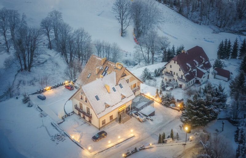 Nocne zdjęcie ośnieżonego zbocza góry, u podnóża której znajdują się oświetlone ciepłym światłem budynki.