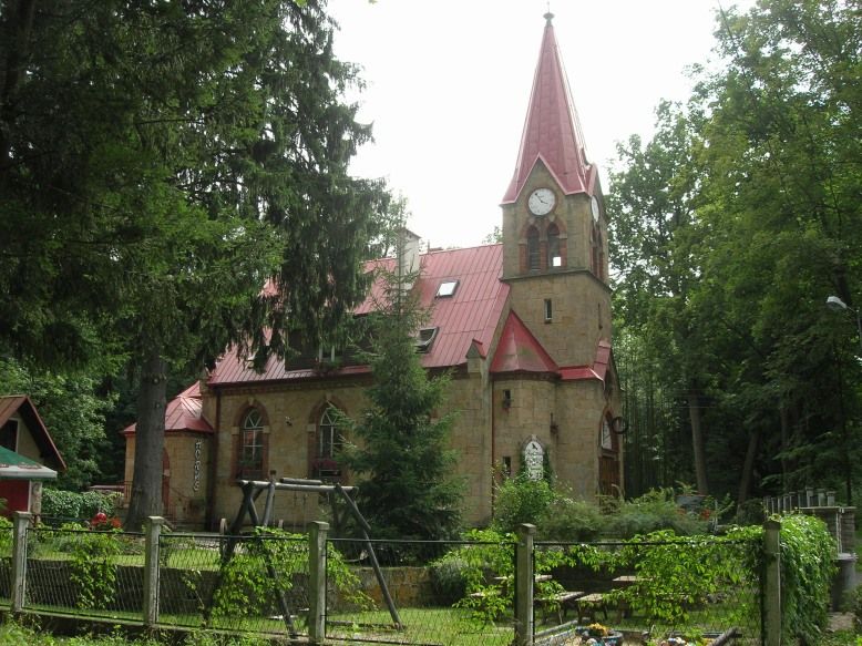 Schowany wśród gęstej roślinności budynek kościoła ze strzelistą wieżą z czerwonym dachem. Przed kościołem stoi huśtawka i ławki.