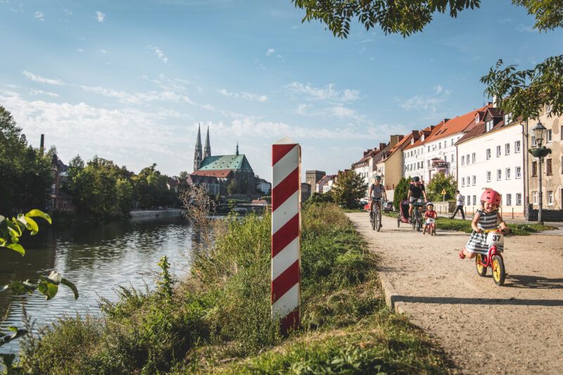 Widok rzekę oraz nadbrzeże, po którym jedzie rodzina na rowerach przy słonecznej pogodzie na tle błękitnego nieba.