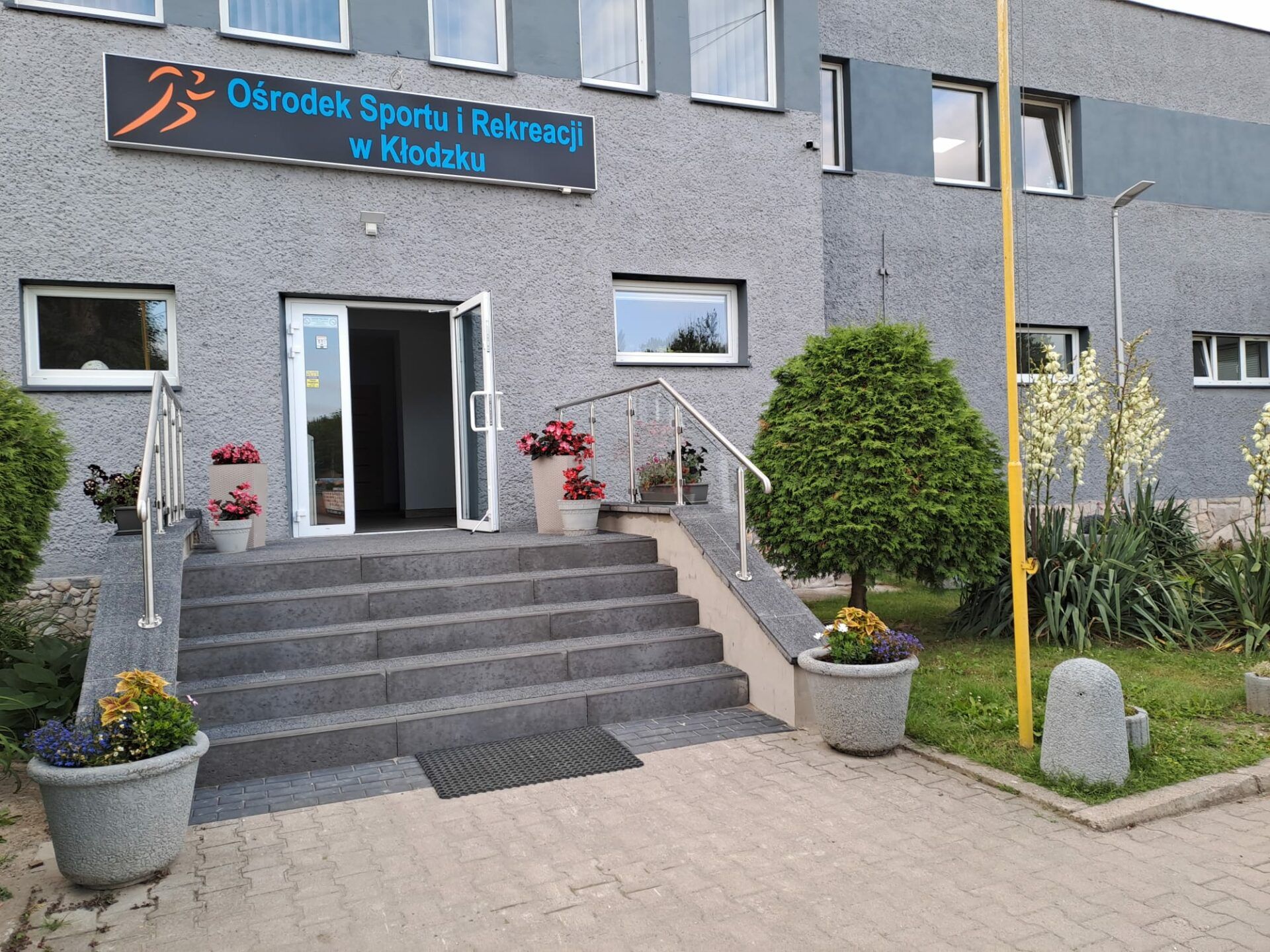 Wejście do szarego, dwupiętrowego budynku, do którego prowadzą schody, zabezpieczone poręczą. Przed wejściem znajdują się donice z nasadzeniami oraz kawałek drobnej ogrodowej zieleni.