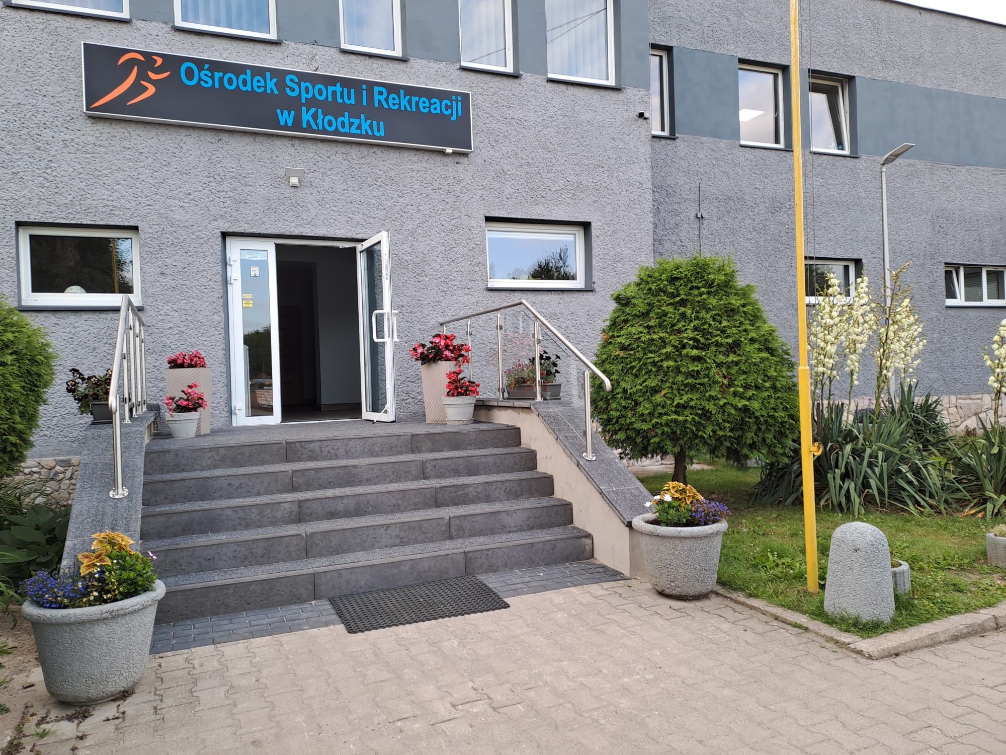 Wejście do szarego, dwupiętrowego budynku, do którego prowadzą schody, zabezpieczone poręczą. Przed wejściem znajdują się donice z nasadzeniami oraz kawałek drobnej ogrodowej zieleni.