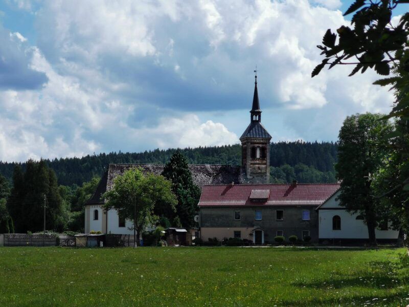 Kamiennny budynek kościoła z jedną wieżą, wśród pól i budynków gospodarczych.
