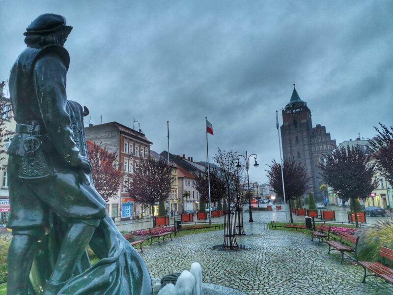 Widok zza pleców pomnika mężczyzny, zwróconego twarzą w stronę Baszty Tkaczy - budynku z wysoką wieżą.
