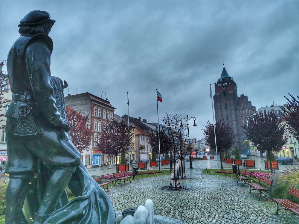 Widok zza pleców pomnika mężczyzny, zwróconego twarzą w stronę Baszty Tkaczy - budynku z wysoką wieżą.