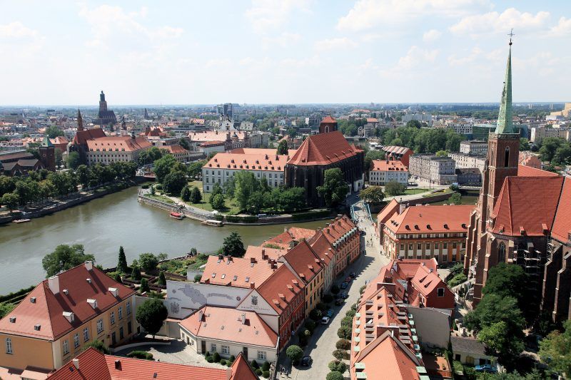 Panorama miasta z zabytkowymi budynkami pokrytymi czerwonymi dachówkami,  znajduje się wyspa i przepływająca rzeka  Odra