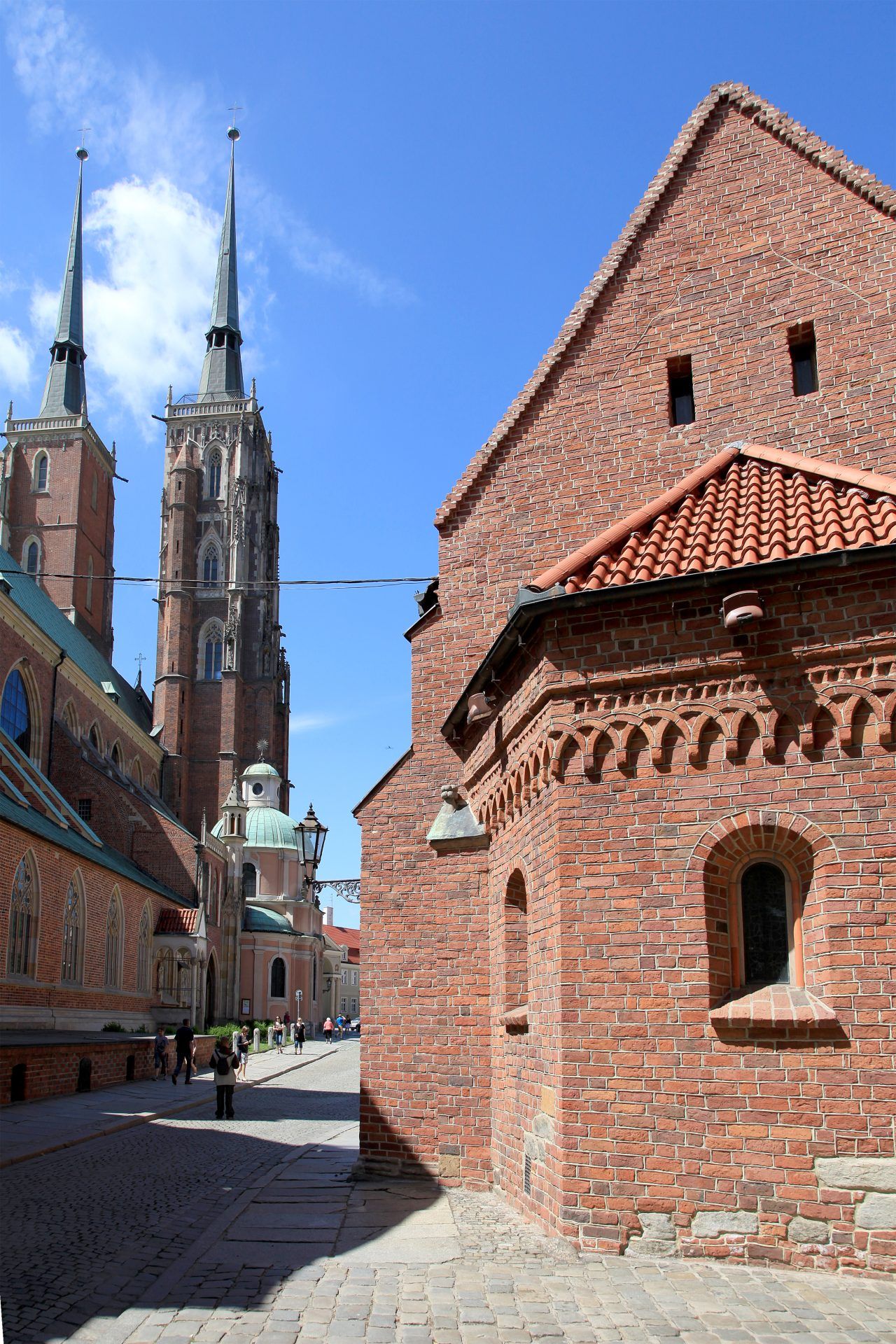 Przejście między dwoma zabytkowymi  zbudowanymi z cegły budynkami z widokiem na wieżę katedry