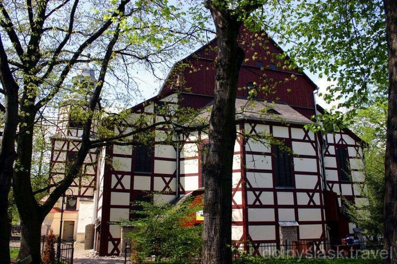 Zewnętrzna biała  ściana w kształcie muru pruskiego- drewnianej konstrukcji kościoła otoczonego wysokimi drzewami
