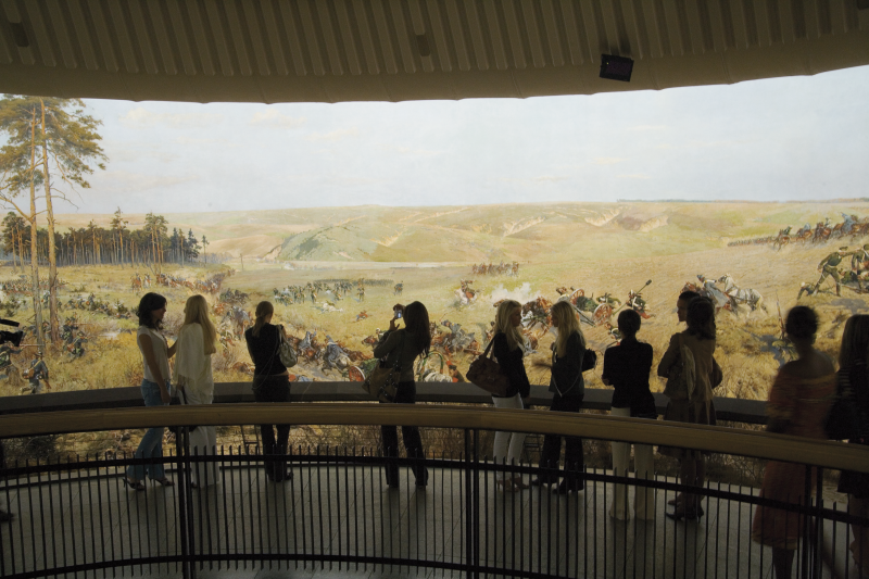 Wnętrze wystawy w Panoramie Racławickiej z widocznym obrazem przedstawiającym bitwę oraz turystami spacerującymi na pierwszym planie