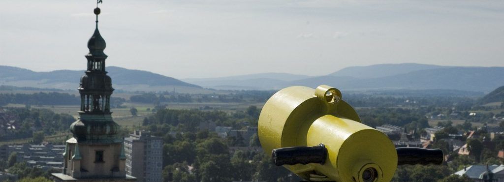 Panoramiczne zdjęcie z widokiem na tereny powiatu Kłodzkiego. W oddali widać zabudowania oraz otaczające powiat wzniesienia górskie 