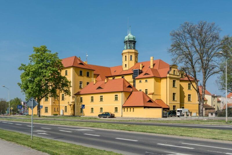 Ceglana, czerwonawo-brązowa budowla z białymi detalami wokół okien i bram, wyróżniająca się wieżą w kolorze czerwonej cegły.