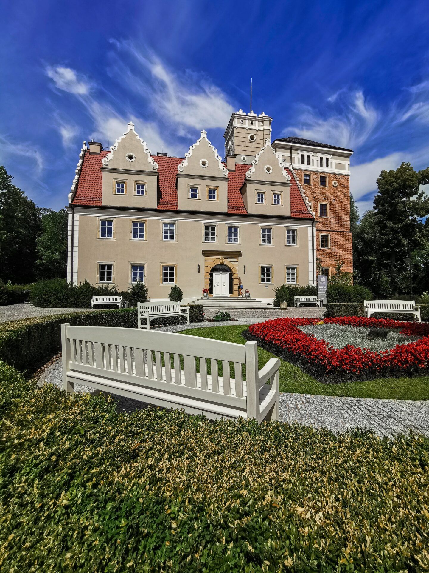 Widok na zamek z jasną elewacją, czerwonym dachem, przed zamkiem zadbany trawnik z kwiatami, na pierwszym planie biała ławka.