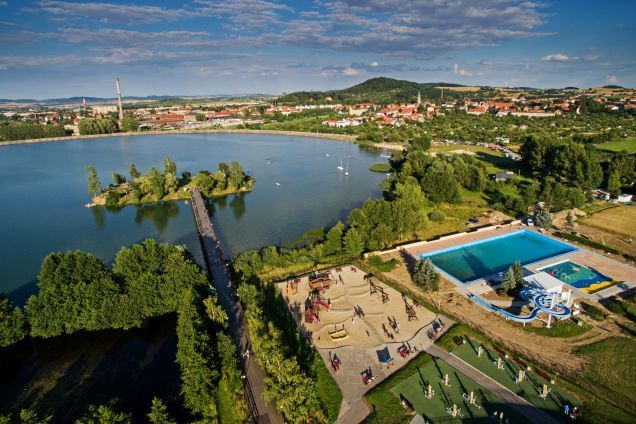 Widok z lotu ptaka na jezioro, otoczone zielenią, domkami, na środku jeziora wysepka.