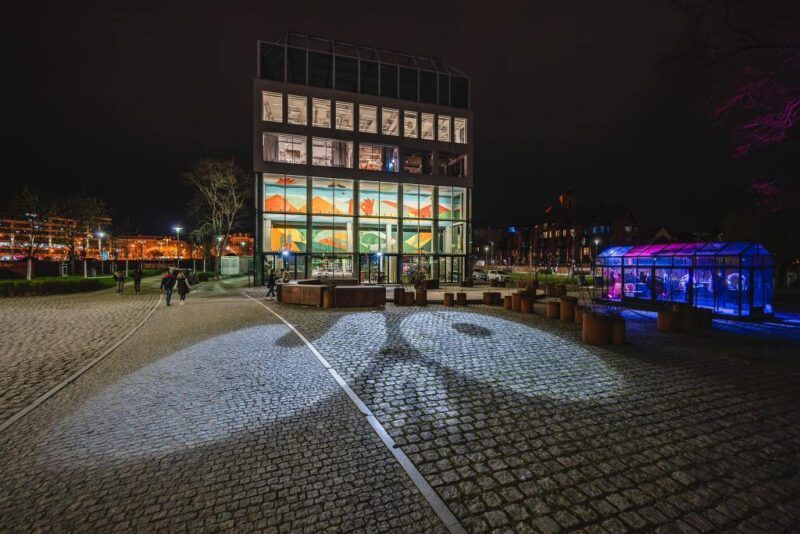 kwadratowy budynek który jest restauracją. jest on cały przeszklony, zdjęcie zostało wykonane w nocy przez co widać wnętrze i kolorowe obrazy znajdujące się w nim. budynek rowniez 