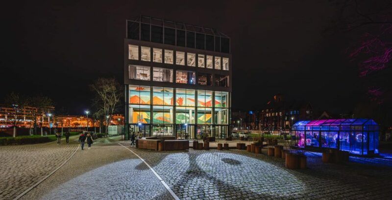 prostokątny budynek który jest restauracją. jest on cały przeszklony, zdjęcie zostało wykonane w nocy przez co widać wnętrze i w nim kolorowy mural znajdujące się w nim. oprócz budynku zdjęcie obejmuje rownież część wyspy słodowej we wrocławiu